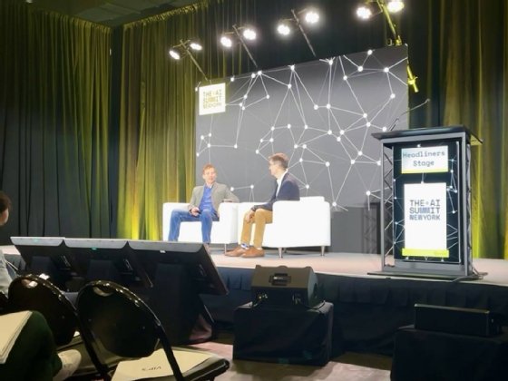 Two people sit on a stage engaged in conversation. The phrase 'The AI Summit New York' appears on a background behind them.