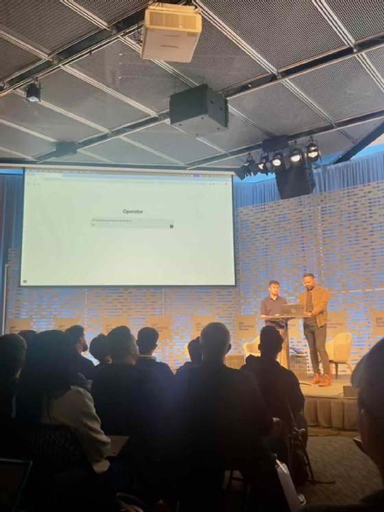 Audience watching a demo at EmTech AI, with two presenters onstage in front of a large screen displaying the Operator agent interface.