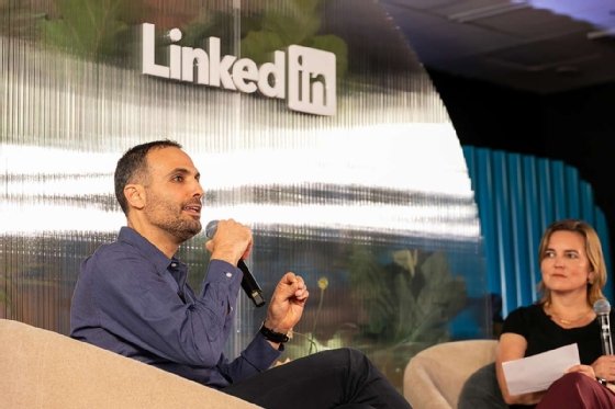 A man speaks into a microphone on a LinkedIn stage while a woman with a microphone and notes listens.